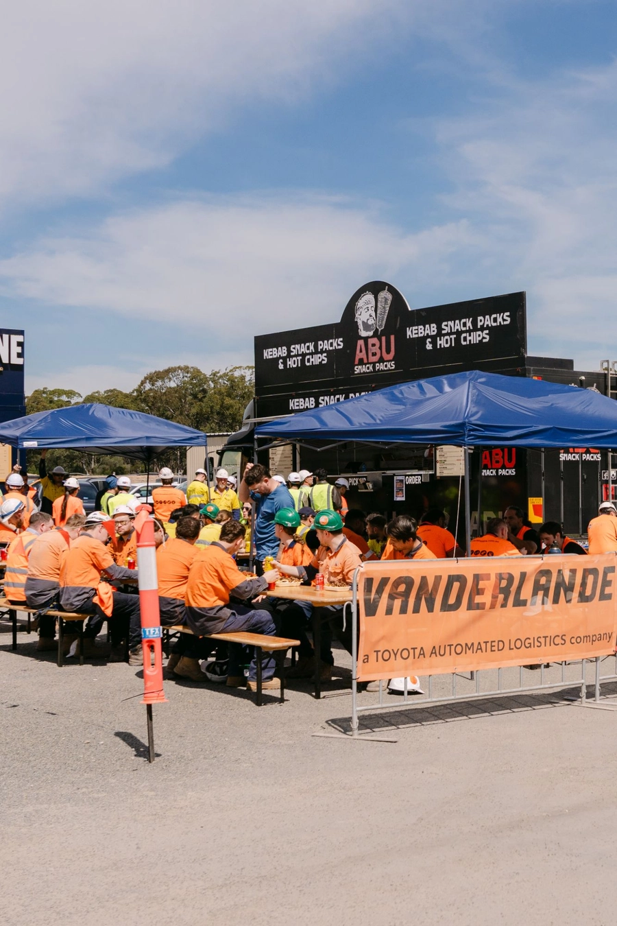 Workers eating snack packs at long tables in front of the Abu Snack Packs truck, beside a Vanderlande banner, at the Moorebank site lunch