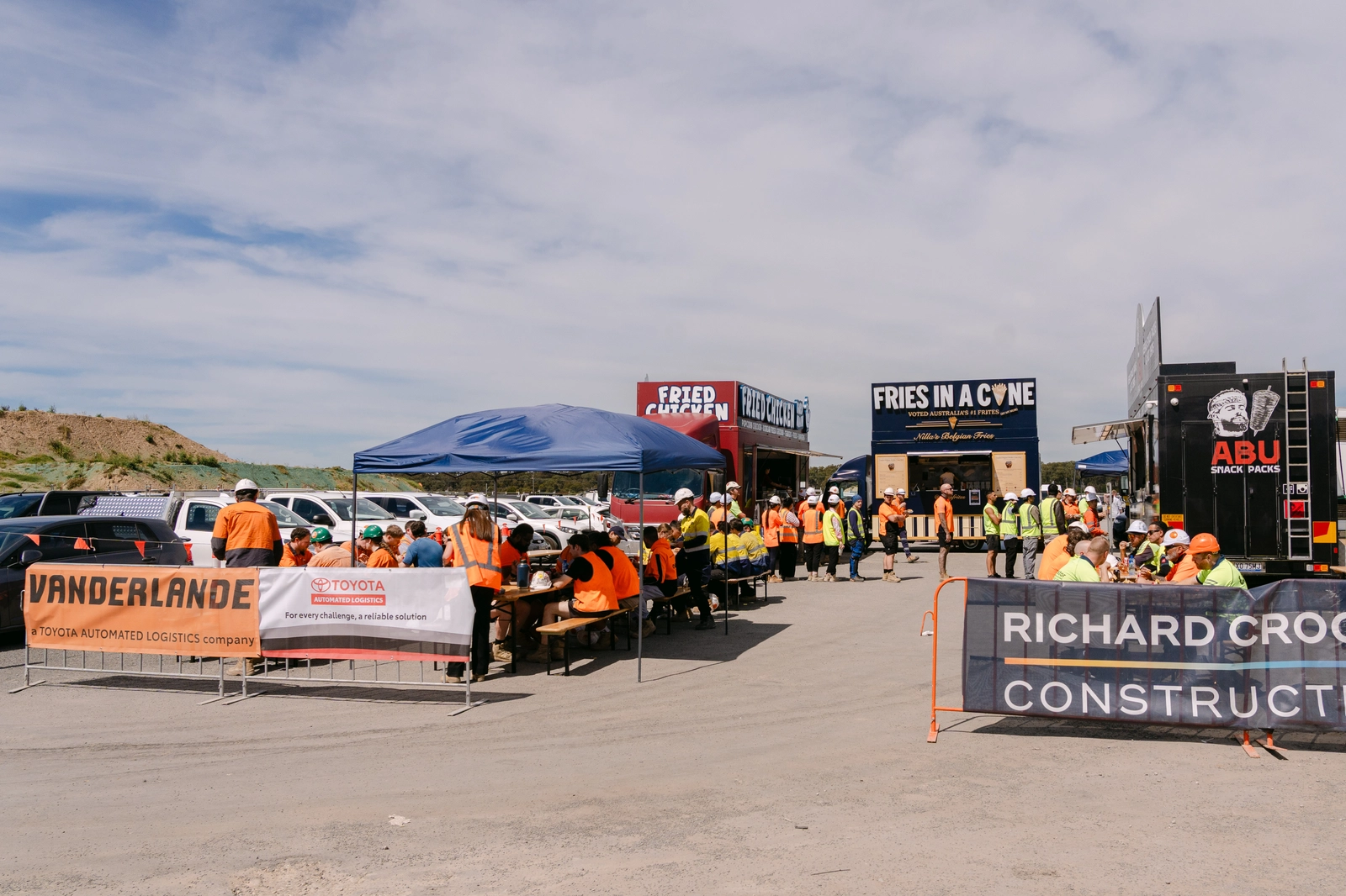 Vanderlande and Richard Crookes Constructions banners in front of three Food Hub trucks, with site workers eating at long tables under a gazebo in Moorebank